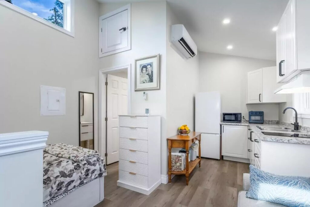 Bright, compact studio with a bed, white cabinetry, a small kitchen, mini fridge, microwave, and wall art. Wood floors and natural light give a cozy, modern feel. A fruit bowl sits on a wooden table.