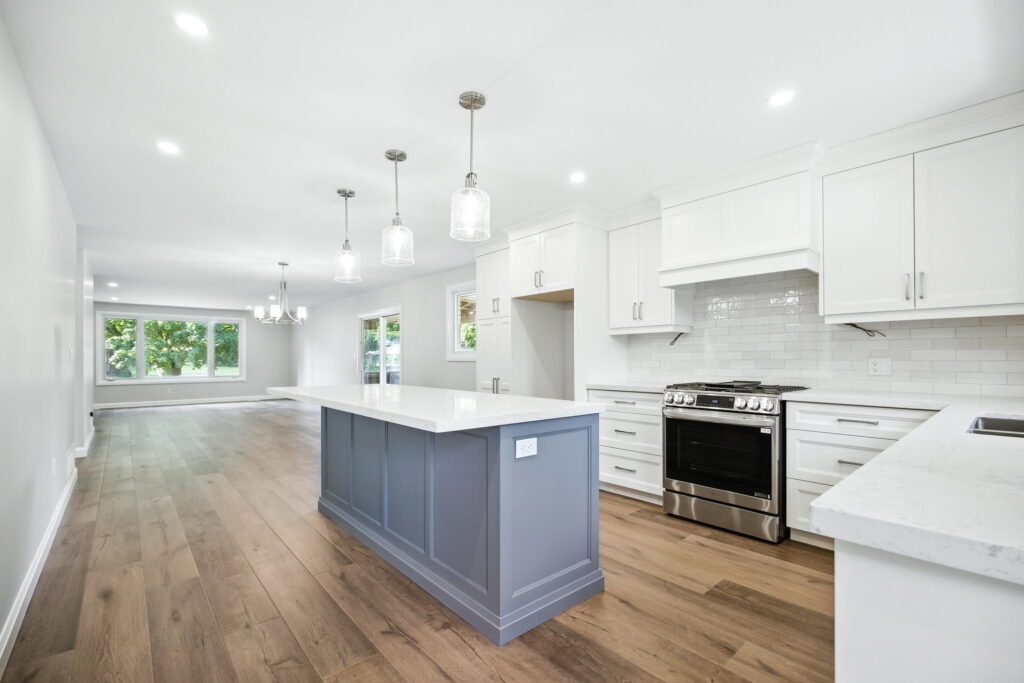 Bright, modern kitchen with white cabinets, stainless steel appliances, a large gray island with a marble countertop, pendant lights, and wood flooring, opening into a spacious living area with large windows.