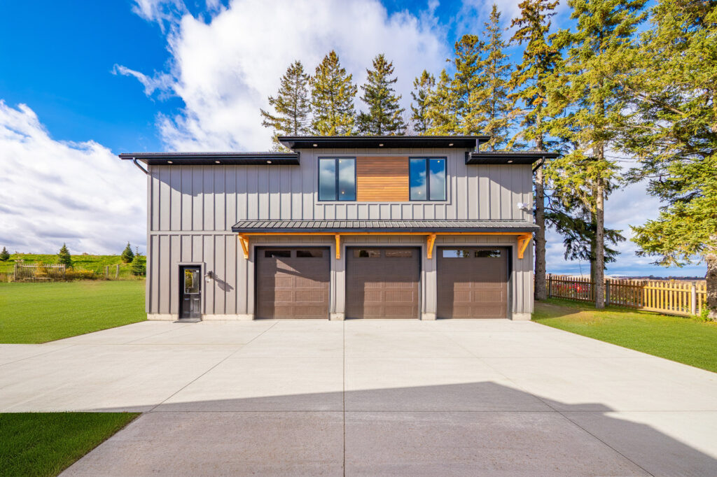 A modern two-story house with gray siding, three large garage doors, a wide concrete driveway, and tall trees behind it under a partly cloudy sky.
