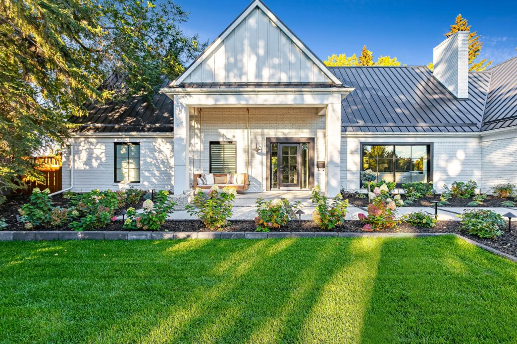 Modern white house with black roof, large windows, and a front porch with a swing. The yard features green grass, landscaped flower beds, and trees with sunlight casting long shadows.