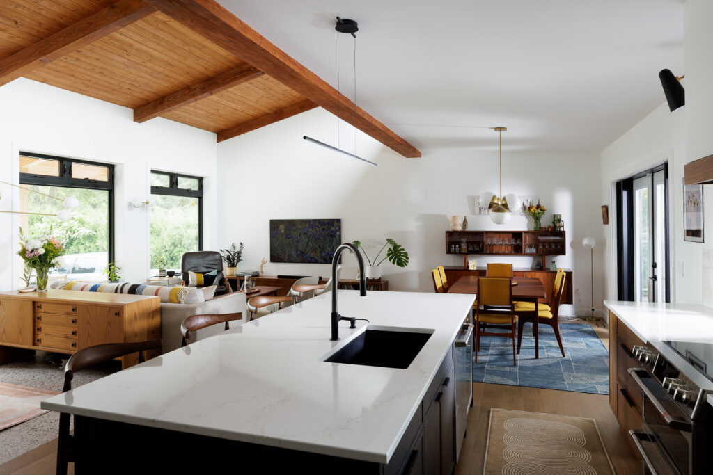 Modern open-concept kitchen and dining area with wooden beams, white countertops, black fixtures, mid-century furniture, large windows, and a mix of natural and artificial lighting.