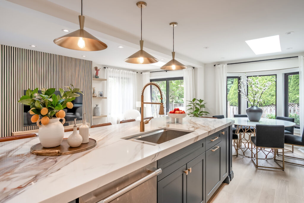 Modern kitchen with a marble island, gold pendant lights, a sink, decorative vases with lemons, and an open dining area with large windows overlooking greenery.