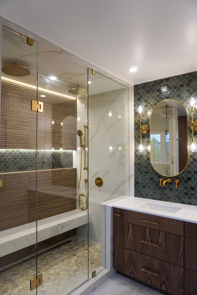 Modern bathroom with a glass-enclosed shower featuring gold fixtures, marble walls, and pebble flooring. The vanity has a white countertop, wood cabinets, and gold accents. Green tile backsplash and decorative lighting are visible.