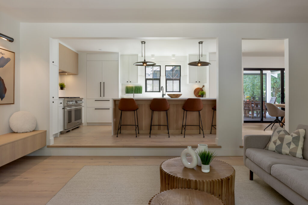 A modern kitchen with light wood cabinets, a large island with four brown barstools, pendant lights, and neutral decor. The kitchen opens to a cozy living area and a dining space visible through glass doors.