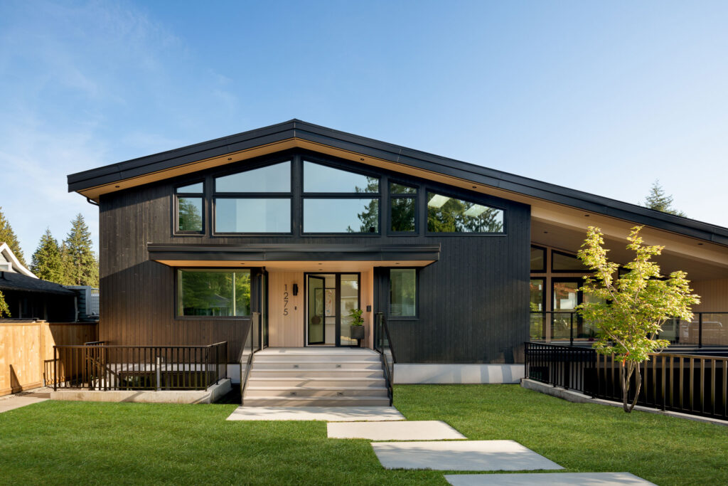 A modern, dark-wood house with large windows, sloped roof, and a covered porch sits on a green lawn with stone steps leading to the entrance. Trees and a blue sky are visible in the background.