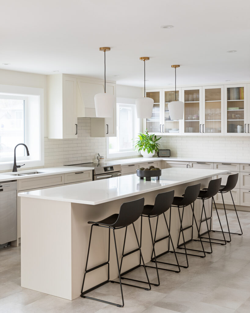 Modern kitchen with a large white island, five black bar stools, white cabinets, stainless steel appliances, three pendant lights, a window above the sink, and a potted plant near glass-fronted upper cabinets.