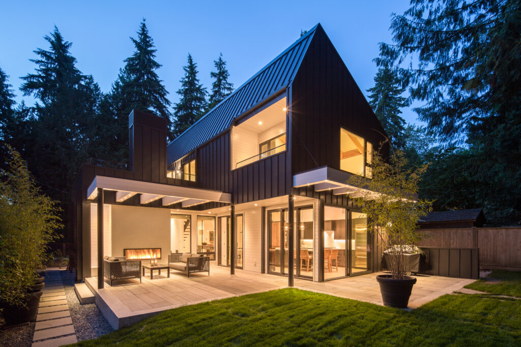 Modern two-story house with large glass windows and doors, dark metal roof and siding, spacious patio with outdoor seating, warm interior lighting, surrounded by grass, trees, and a wooden fence at dusk.