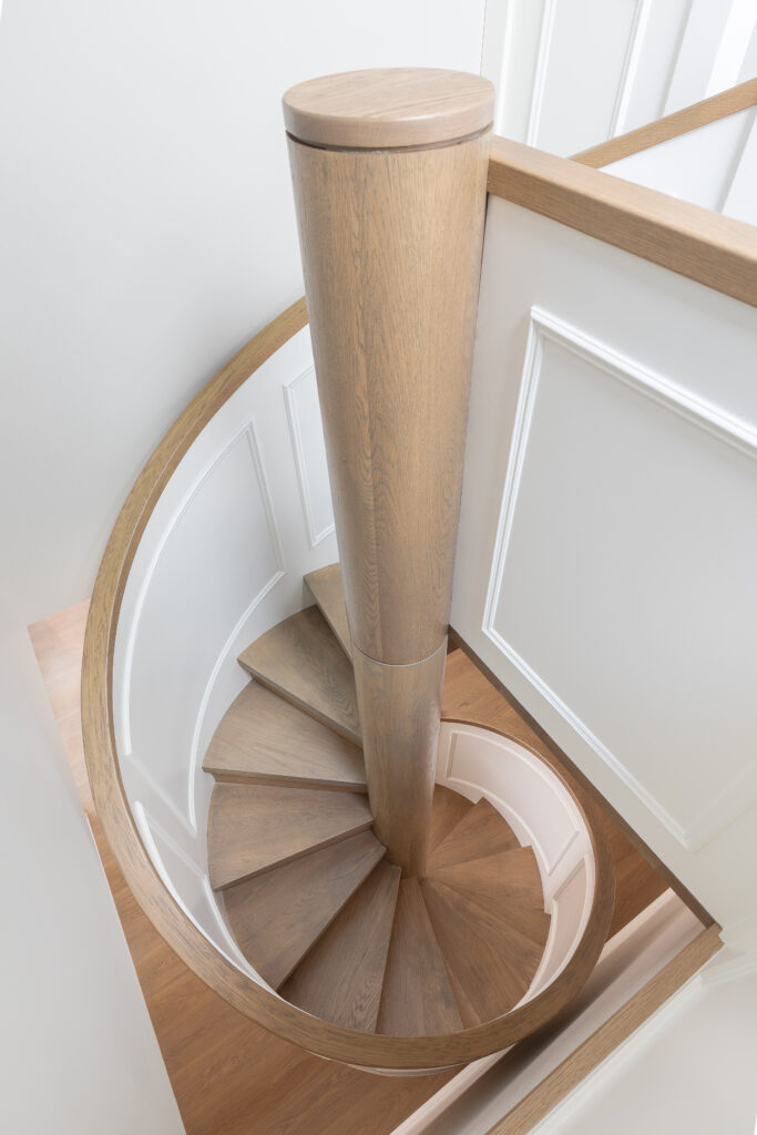 A top-down view of a modern, spiral wooden staircase with white paneling on the walls and a central column, leading down to a light-toned wooden floor.