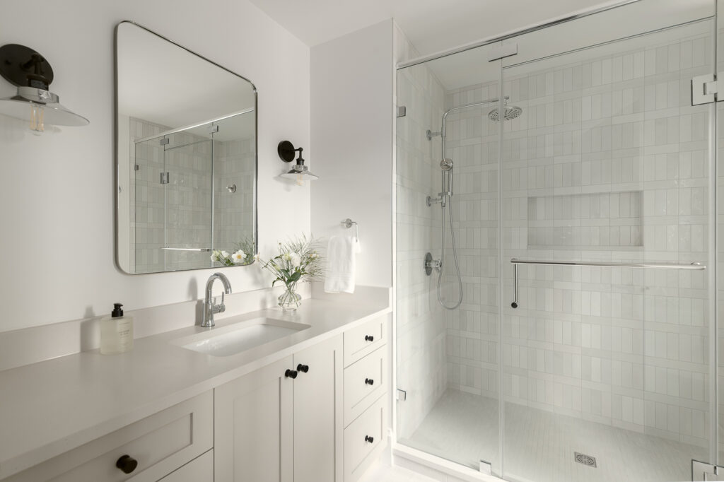 A modern bathroom with a large glass shower, white tile walls, a white vanity with drawers and cabinets, a countertop sink, a wall mirror, and black sconces. A vase with flowers sits next to a hand towel.