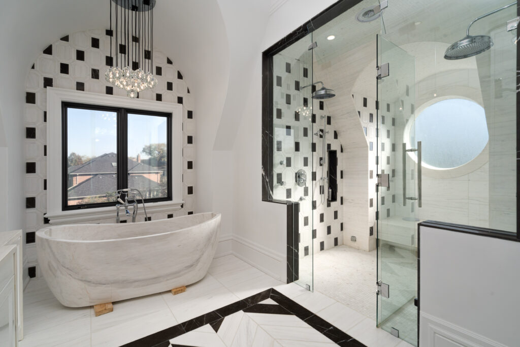 Modern bathroom with a freestanding marble bathtub, black-and-white tiled walls, large window, glass-enclosed shower with dual showerheads, and a chandelier overhead. Natural light fills the space.