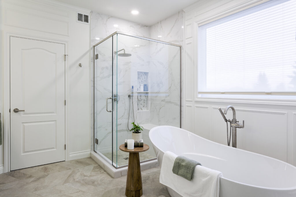 Modern bathroom with a glass-enclosed shower, a white freestanding tub, a small wooden stool with a plant and candle, marble-patterned walls, and a large window with blinds.