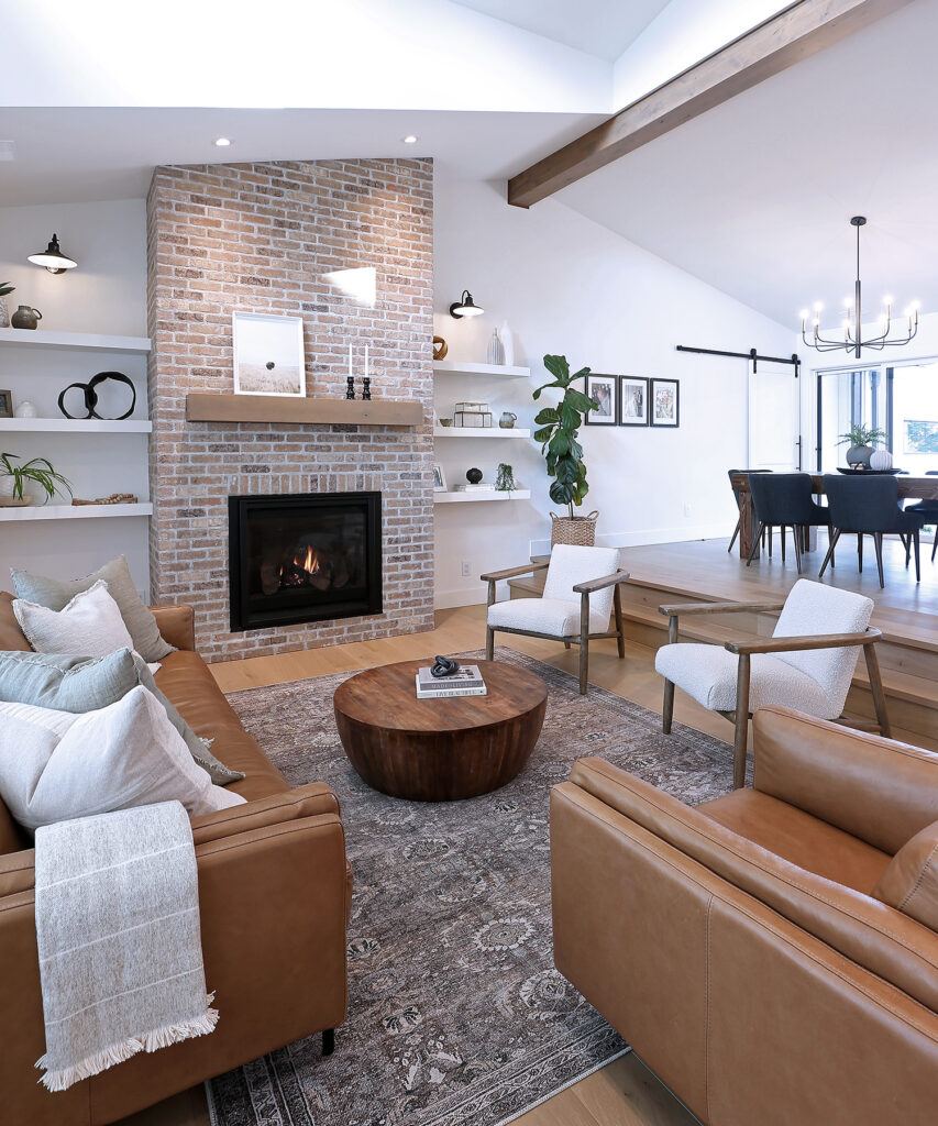 A modern living room with tan leather sofas, a round wooden coffee table, a brick fireplace, built-in shelves with decor, and a dining area in the background. The space is bright with natural light and greenery.