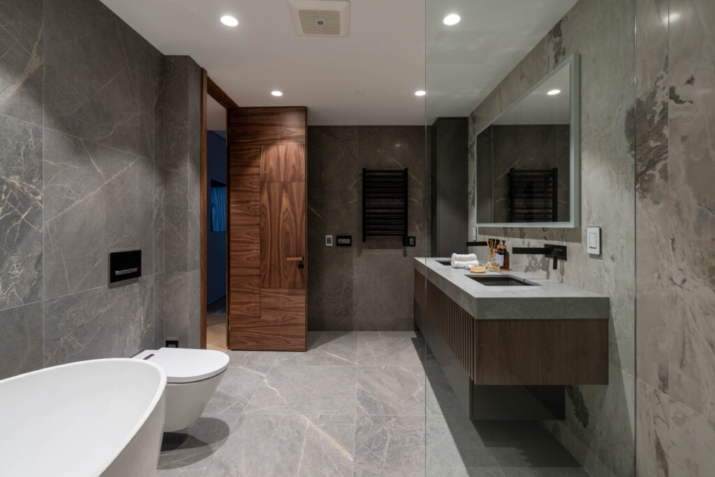 A modern bathroom with gray marble walls and floor, a freestanding white bathtub, wall-mounted toilet, floating vanity with a large mirror, and wooden door. The space is well-lit with recessed ceiling lights.
