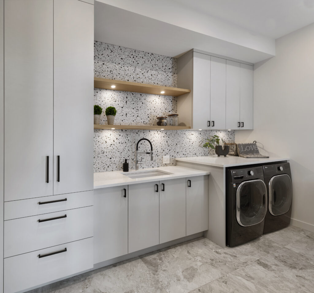 Modern laundry room with light gray cabinets, black washer and dryer, terrazzo backsplash, open wooden shelf with plants and decor, built-in sink, and Love sign on the counter. Marble floor tiles.