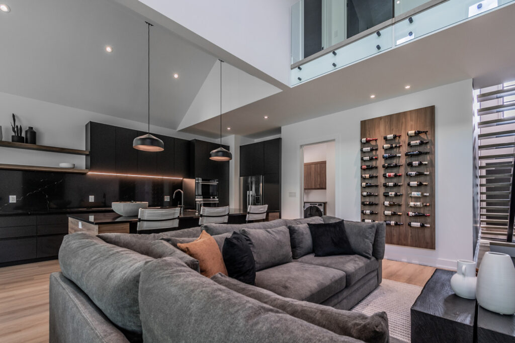 Modern open-concept living space with a gray sectional sofa, dark kitchen cabinets, pendant lights, a large island with bar stools, and a wall-mounted wine rack; staircase and mezzanine visible.