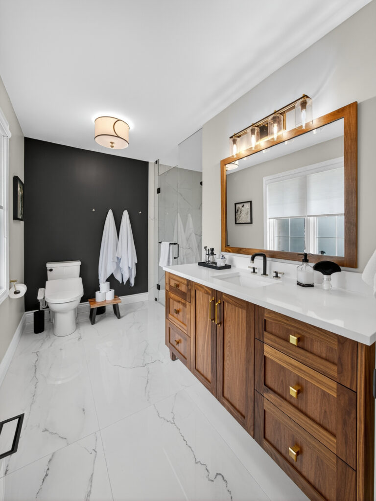 Modern bathroom with a large wooden vanity, double sinks, brass fixtures, white countertops, a wall-mounted mirror, a toilet, towels hanging on a dark accent wall, and a glass shower enclosure on glossy white tile floors.