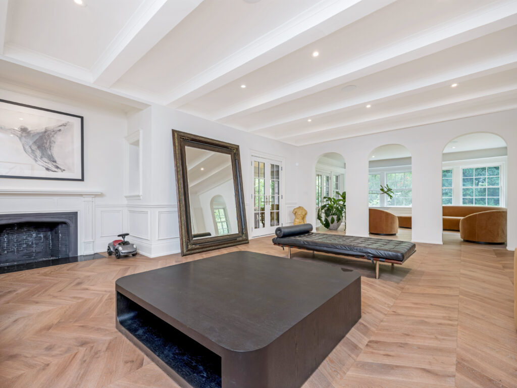 Spacious, modern living room with a large black coffee table, floor mirror, fireplace, and minimalist decor. Arched doorways and tall windows allow natural light to fill the room with wooden herringbone flooring.