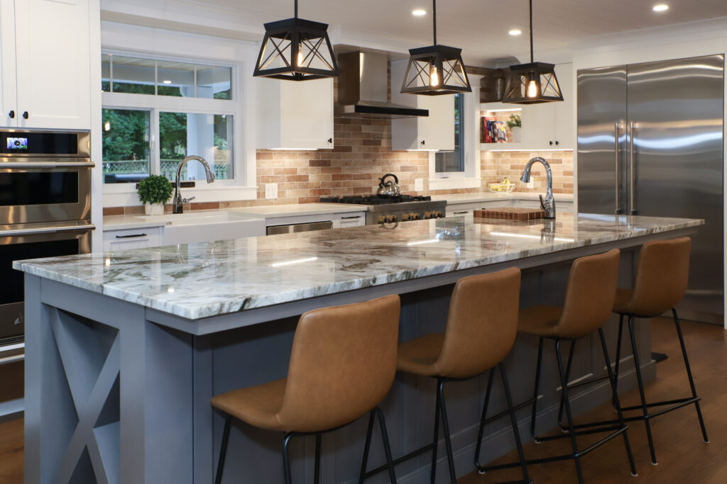Modern kitchen with a large marble island, four brown leather bar stools, stainless steel appliances, brick backsplash, white cabinets, and pendant lights hanging above the counter.