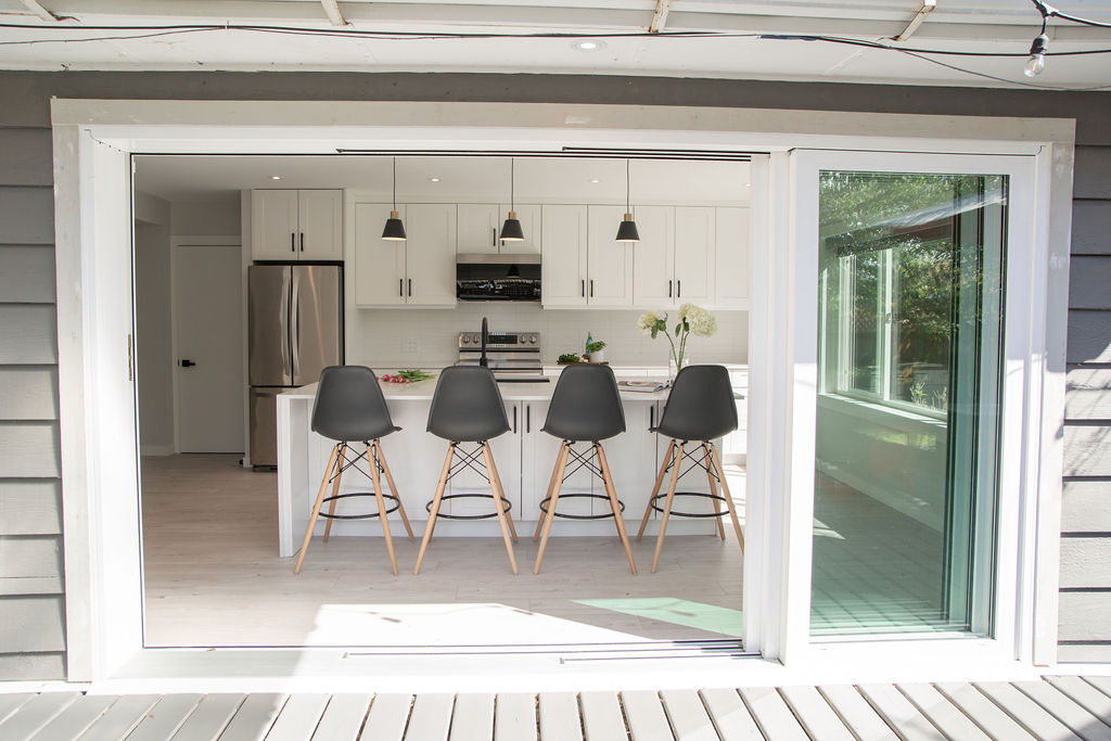 A modern kitchen with white cabinets, a center island, and four black barstools is viewed through large glass sliding doors that open onto a deck. Pendant lights hang above the island.