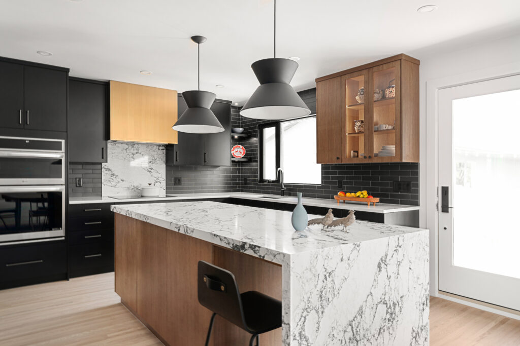 Modern kitchen with black and wood cabinets, marble countertops, black tile backsplash, two black pendant lights, a built-in oven, a bar stool, and a vase and fruit bowl on the island near a glass door.