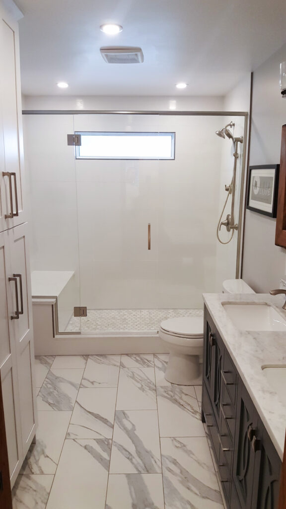 Modern bathroom with marble tile floor, glass-enclosed shower with a built-in bench, rain showerhead, vanity with dual sinks and white countertops, and bright overhead lighting.