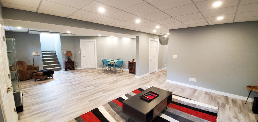A spacious, modern basement with light gray walls and wood-look flooring. There’s a sitting area with chairs, a small dining area with blue chairs, a coffee table with a rug, and stairs leading up. Ceiling lights illuminate the room.