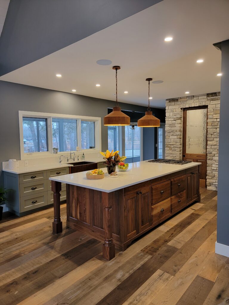 Modern kitchen with a large wooden island, white countertops, pendant lights, stainless steel sink, fruit bowl, and flowers. The room features wood flooring, gray walls, and a stone accent wall with a wooden door.