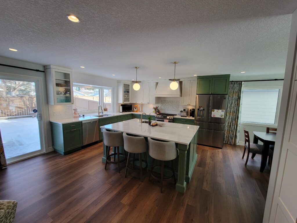 A modern kitchen with green cabinetry, a large white island with four gray barstools, stainless steel appliances, wood flooring, pendant lights, and a dining area visible in the background.