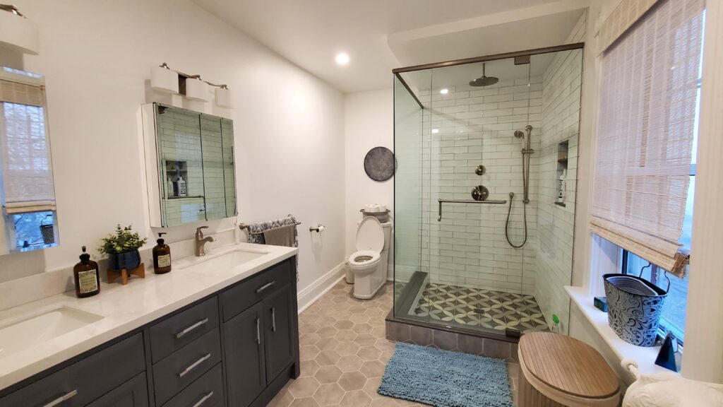 Modern bathroom with a double-sink vanity, large mirror, walk-in glass shower, toilet, gray cabinets, hexagonal floor tiles, and a window with blinds. Blue rug and decorative items add a cozy touch.