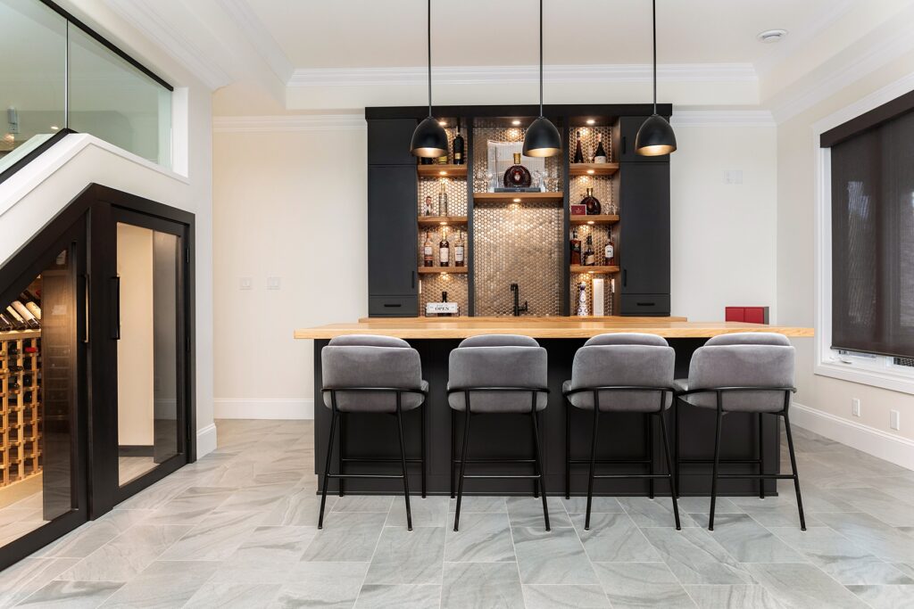 A modern home bar with a wooden countertop, four gray upholstered bar stools, black cabinets, shelves with bottles and glasses, three black pendant lights, and a wine cellar visible under a staircase.