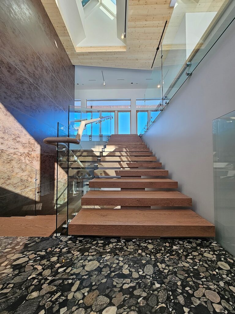 A modern staircase with floating wooden steps, glass railings, and a pebble-patterned floor leads up to large windows that allow natural light to fill the open, airy space.