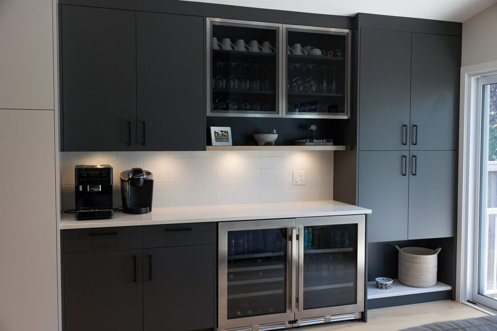 Modern kitchen coffee and beverage station with dark cabinets, under-cabinet lighting, a coffee maker, a Keurig machine, glass-front upper cabinets, wine fridges, and a basket on a lower shelf near a sliding glass door.