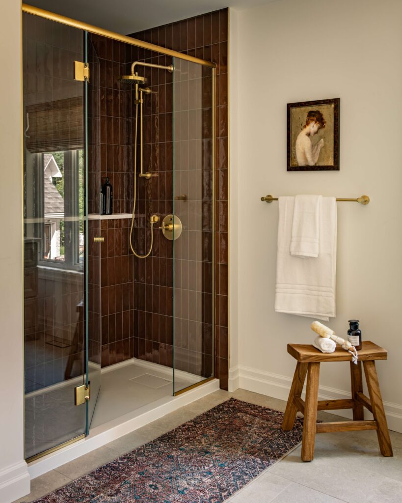 A modern bathroom with a glass shower featuring gold fixtures and brown tiles, a wooden stool with bath items, a white towel on a gold rack, a framed portrait, and a patterned rug on the floor.