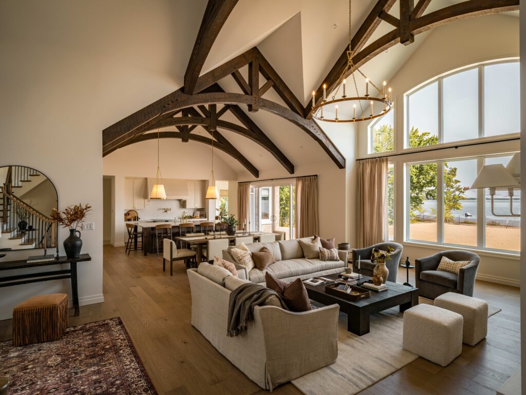 Spacious, elegant living room with exposed dark wood beams, large arched window overlooking water, neutral-toned sofas, a modern chandelier, and open-concept kitchen and dining area in the background.