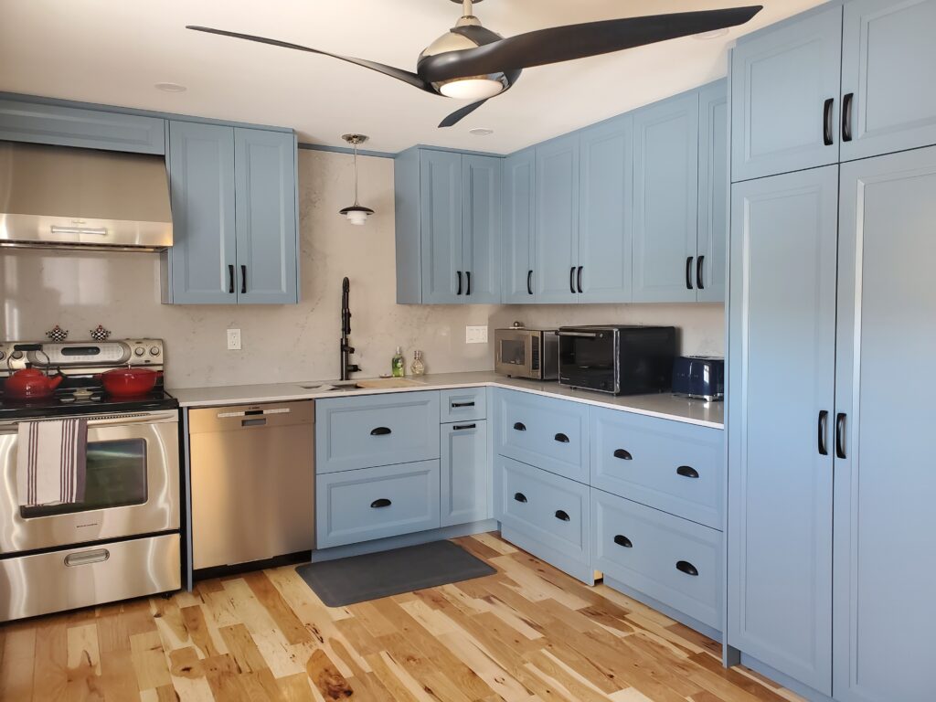 Modern kitchen with light blue cabinets, stainless steel appliances, a black ceiling fan, light wood floors, and various countertop appliances including microwaves and a toaster. Natural light brightens the space.