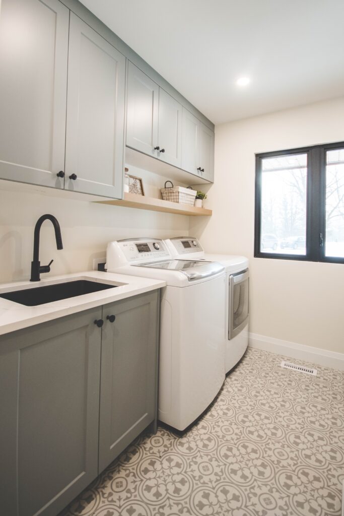 Modern laundry room with gray cabinets, a black sink faucet, a white countertop, a washing machine, a dryer, open wooden shelf with baskets, and a large window. The floor has a decorative gray and white pattern.