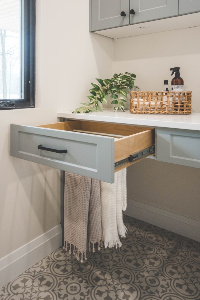 A light blue drawer is open, holding folded beige and white towels. The drawer is beneath a white countertop with a plant, a woven basket, and a spray bottle. The floor has a patterned tile design.