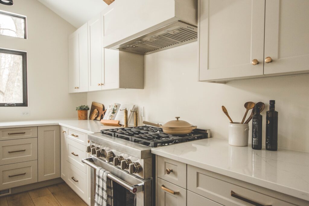 Modern kitchen with white cabinets, a gas stove, beige pot, utensils in a white jar, olive oil bottle, and cutting boards on a white countertop. Natural light comes in through a window on the left.