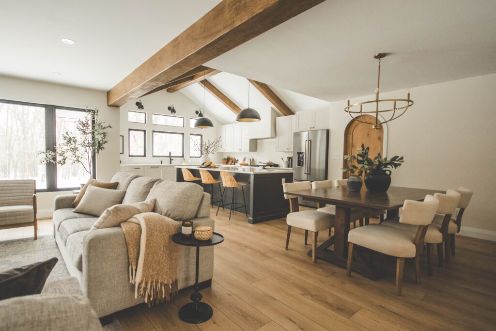 Open-concept living and dining area with light wood floors, beige sofa and chairs, a wooden dining table, a modern kitchen with island seating, and large windows letting in natural light.