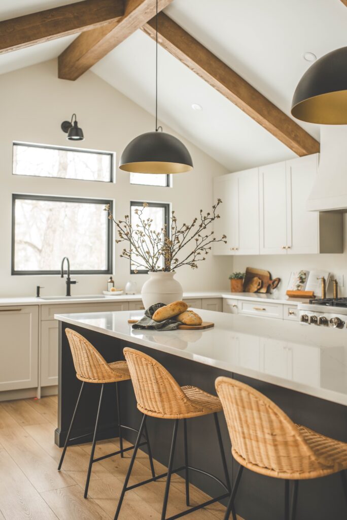 A modern kitchen with white cabinets, wood beams on the ceiling, a central island with wicker bar stools, pendant lights, and a vase with branches. Bread sits on a tray atop the island. Natural light comes through the windows.