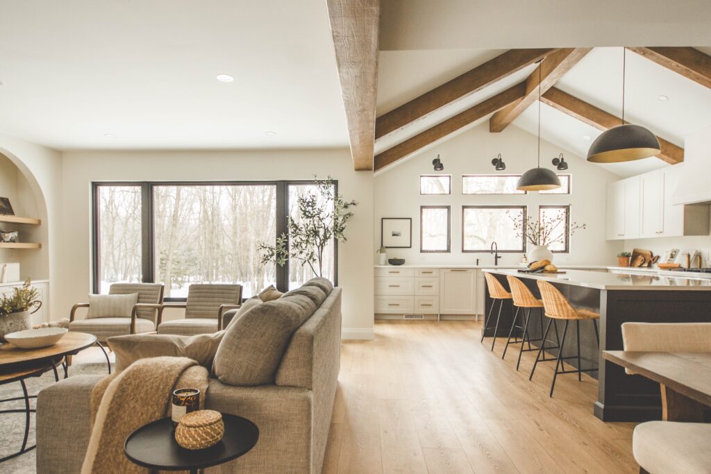 A modern open-concept living room and kitchen with light wood floors, neutral-toned furniture, exposed wooden ceiling beams, large windows, and natural light. Woven bar stools line the kitchen island.