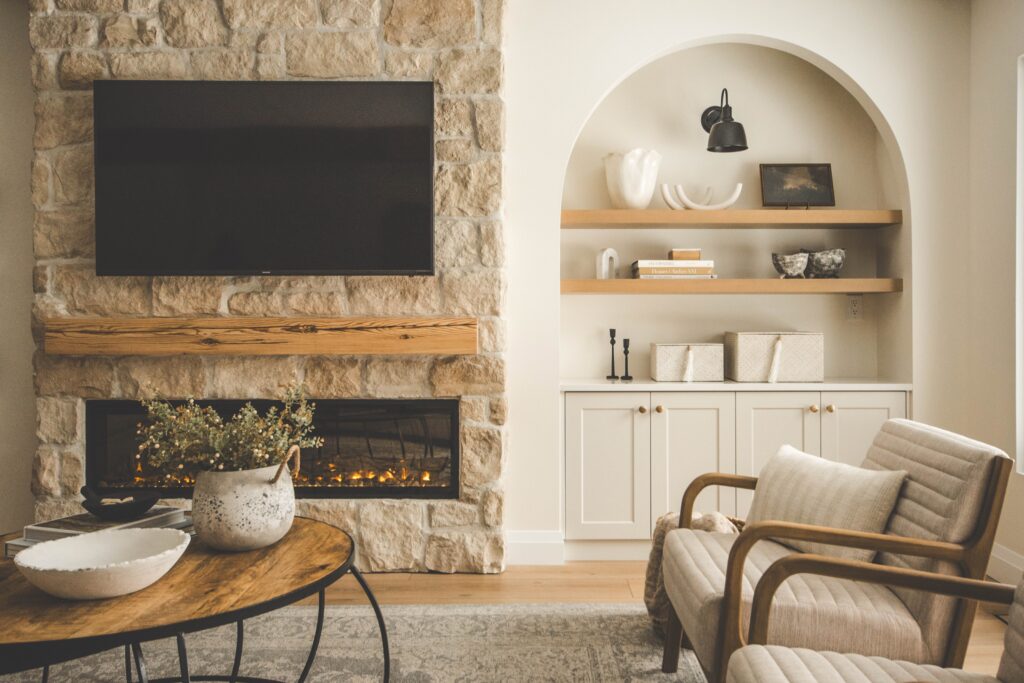 A cozy living room with a stone fireplace, wall-mounted TV, wooden coffee table, armchair, and built-in shelves displaying decor items. Light, neutral tones create a warm, inviting atmosphere.