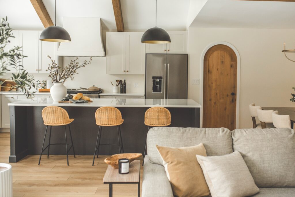 A modern kitchen with a large island, three wicker bar stools, pendant lights, a stainless steel fridge, white cabinets, and wooden accents. A cozy sofa with neutral cushions is in the foreground.