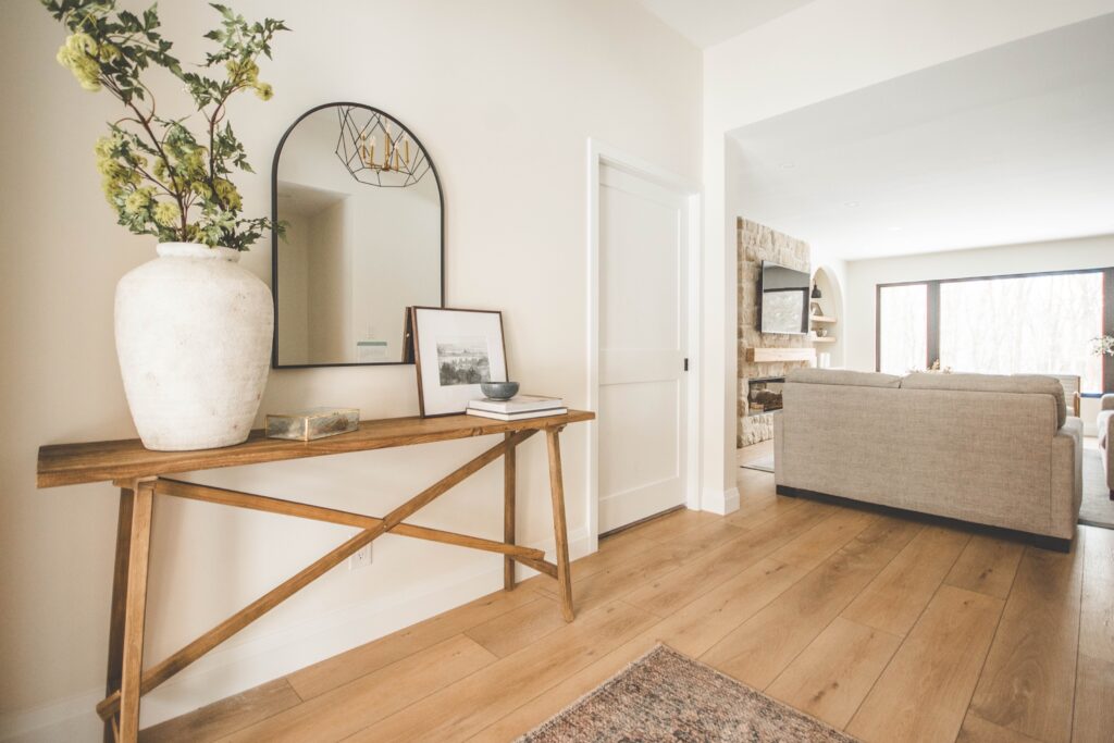 A modern, airy living space with light wood floors, a beige sofa, and a wooden console table topped with a large vase, books, and a framed photo. A round mirror hangs above the console table.