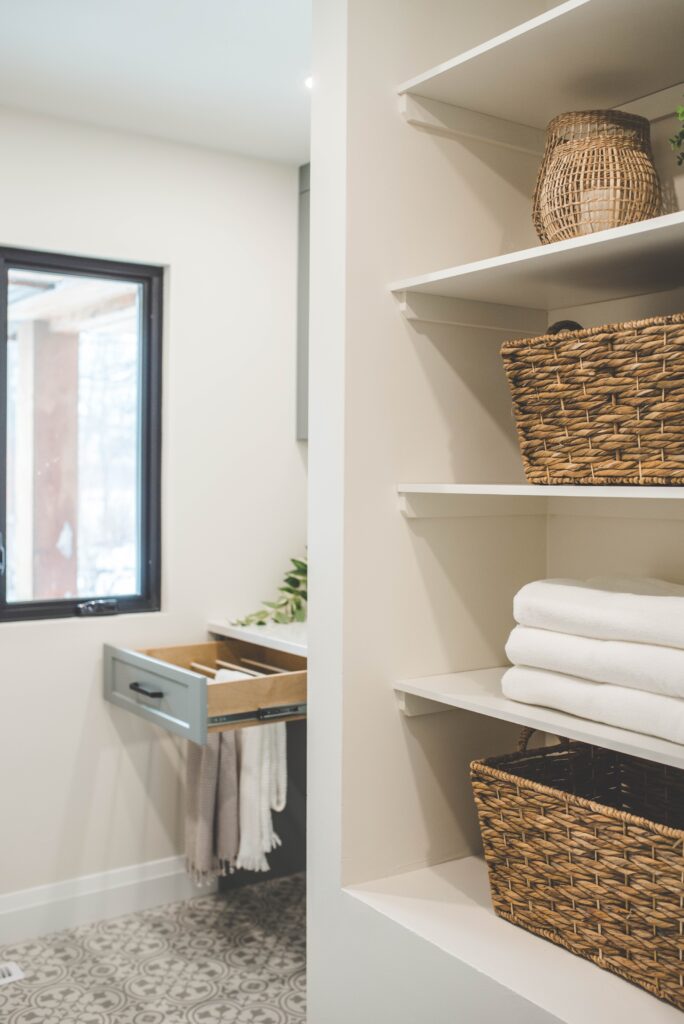 Open shelving with woven baskets and neatly folded white towels in a bright, modern bathroom. A pull-out drawer is open nearby, and a window lets in natural light. The space has a clean, organized feel.