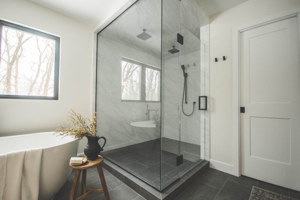 Modern bathroom with a glass-enclosed shower, gray tile floor, freestanding bathtub by a window, and a wooden stool holding a black vase with greenery and a folded towel.