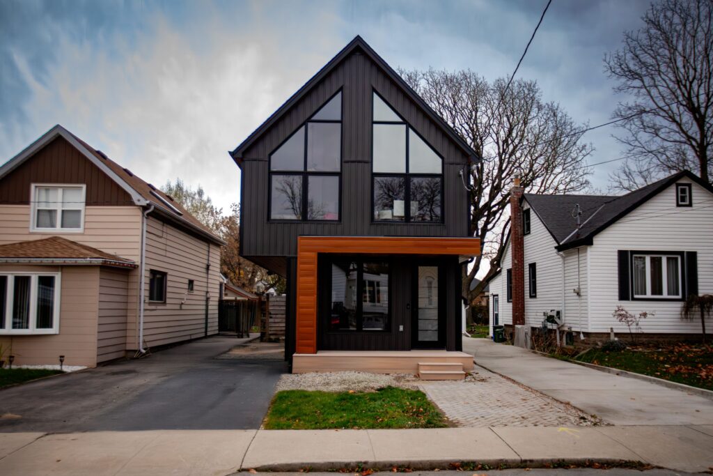 A modern two-story house with dark exterior paneling, large triangular windows, and a wooden accent frame, situated between two older single-story homes, with bare trees and a cloudy sky in the background.