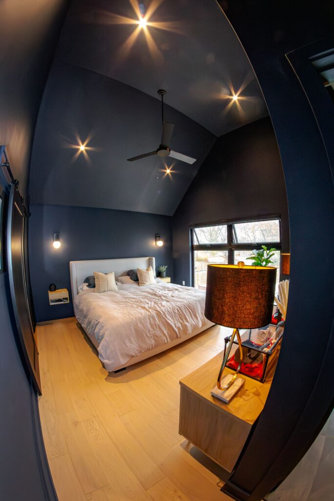 A cozy modern bedroom with dark blue walls, light wood flooring, a bed with white bedding, a ceiling fan, wall sconces, a side table with a lamp, and large windows letting in natural light.