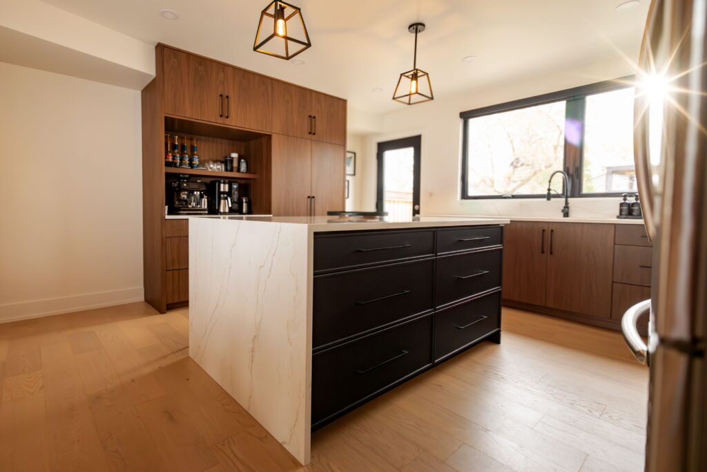 Modern kitchen with wood cabinets, a large black and white marble island, pendant lights, built-in coffee station, light wood floors, and sunlight streaming through a window.