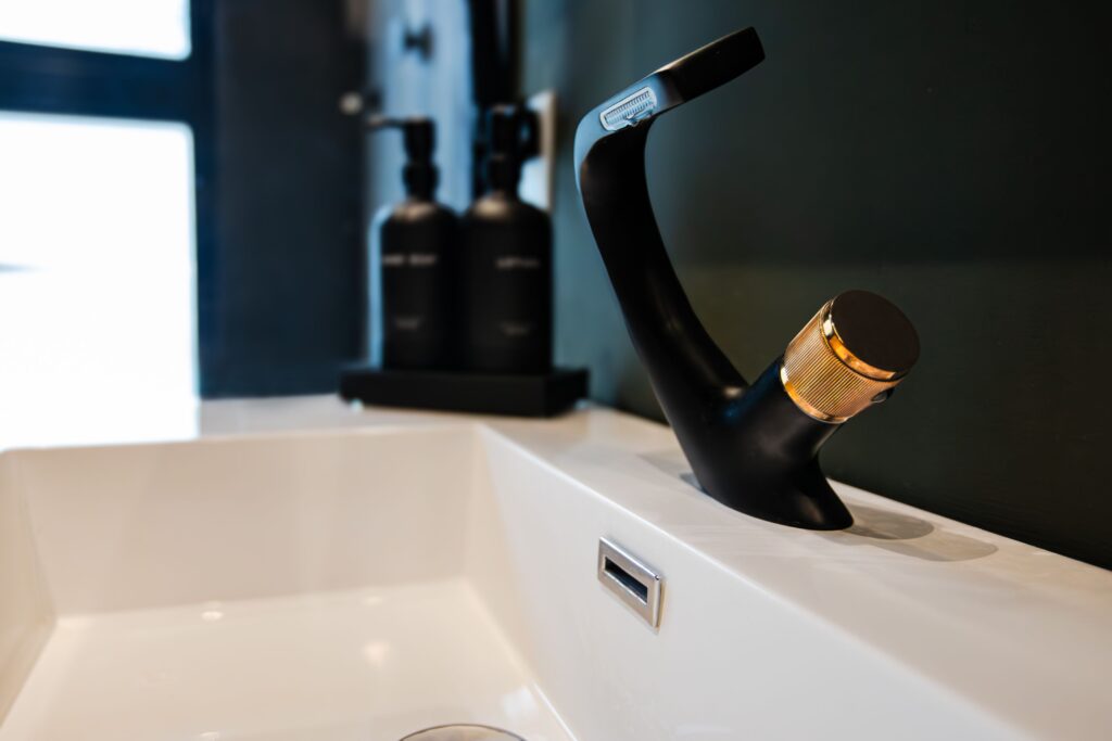 Close-up of a modern black and gold faucet on a white sink, with two matte black soap dispensers in the background against a dark wall.